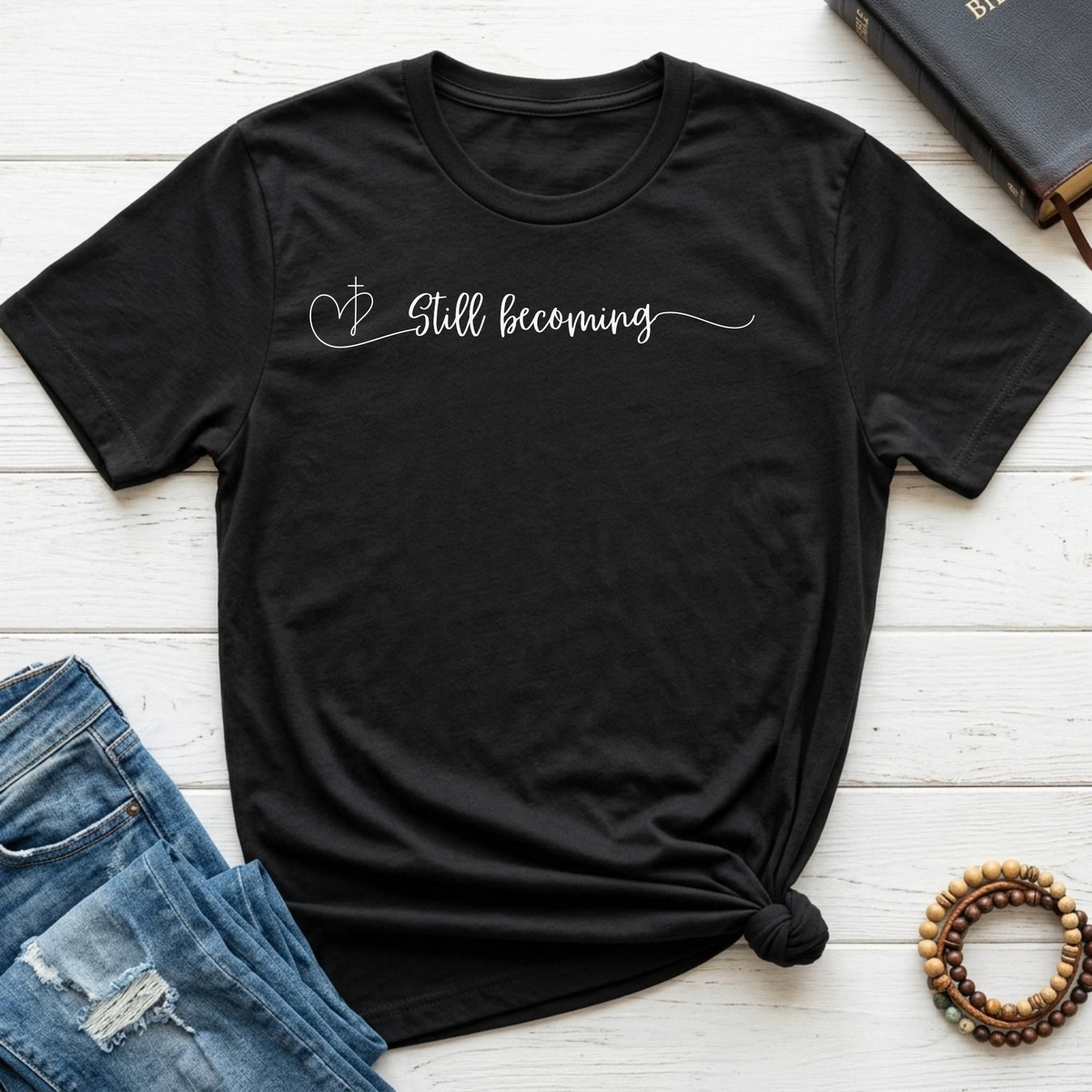 A black short-sleeve t-shirt with 'Still becoming' written in white script across the chest, styled in a flat lay on white wood planks with blue jeans, a book, and a wooden bead bracelet nearby.