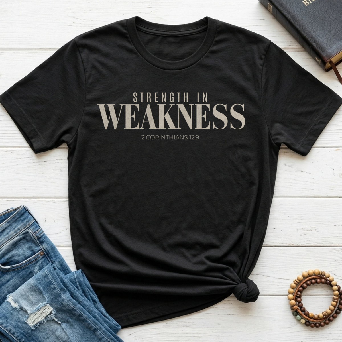 A black short-sleeve graphic T-shirt laid flat on a white wood surface, styled with blue jeans, a book, and a beaded bracelet. The shirt has large white text reading 'STRENGTH IN WEAKNESS' with smaller text '2 CORINTHIANS 12:9' underneath.