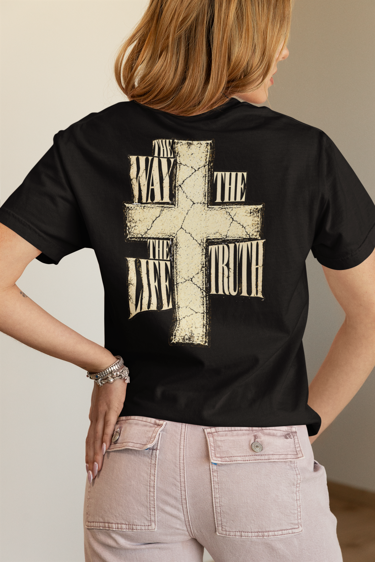 A woman shown from the back wearing a black Christian graphic t-shirt by SeedSowerGraphics with a large distressed cross graphic and the words 'The Way The Life The Truth' printed on it, paired with light pink jeans.