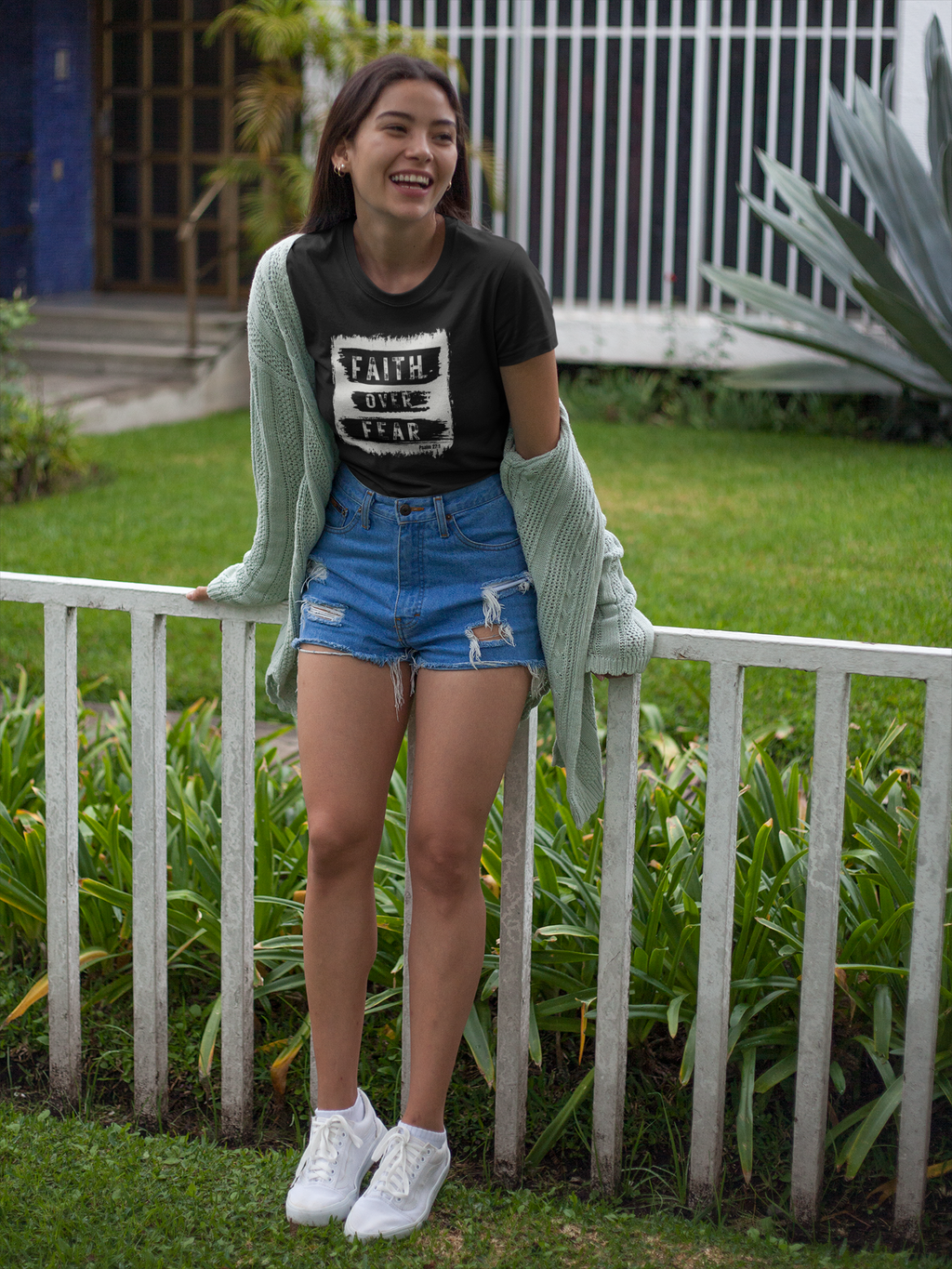 A young woman stands outdoors in a garden setting, leaning against a white picket fence. She is wearing a black Christian graphic t-shirt with 'FAITH over fear" printed on the front, blue denim shorts, a light sage green knit cardigan draped off her shoulders, and white sneakers.