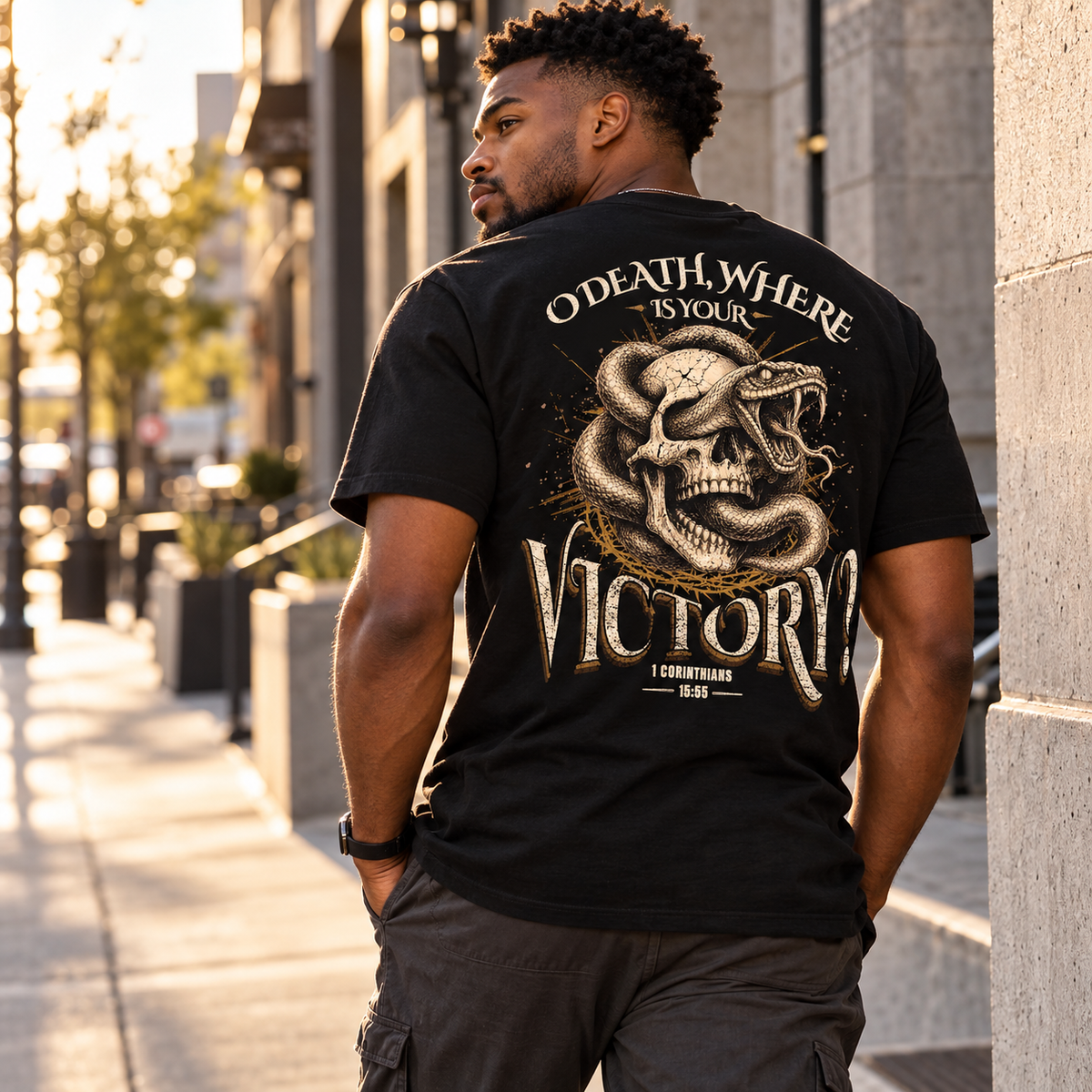 A man standing on a city sidewalk wearing a black graphic T-shirt with a large snake and skull design and the words 'O DEATH, WHERE IS YOUR VICTORY' on the back.