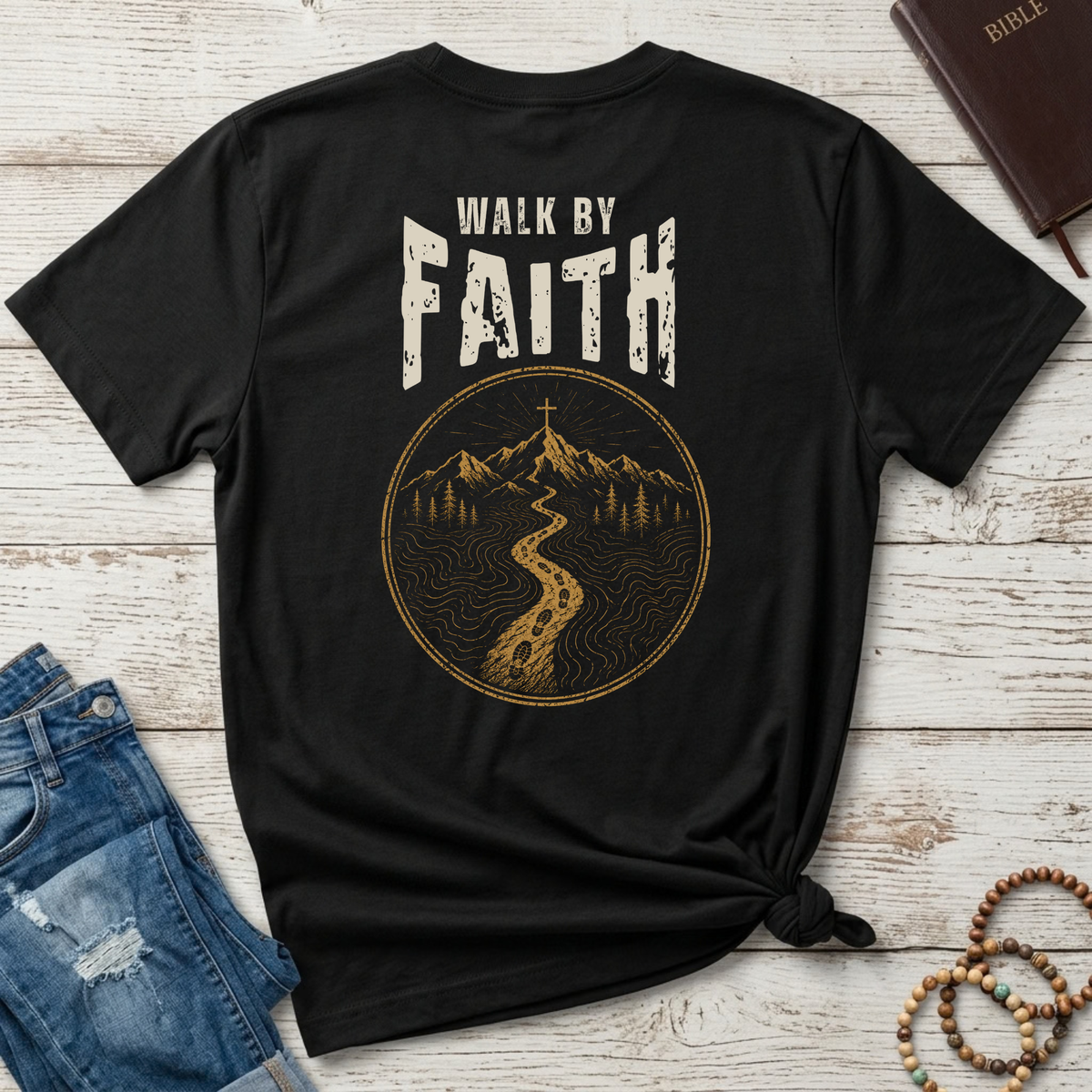A black short-sleeve Christian T-shirt by SeedSowerGraphics laid flat on a whitewashed wood surface, styled with jeans, a Bible, and beaded bracelets. The shirt shows a large distressed graphic on the back with the words “WALK BY FAITH” above a circular mountain-and-path illustration in tan/gold tones.