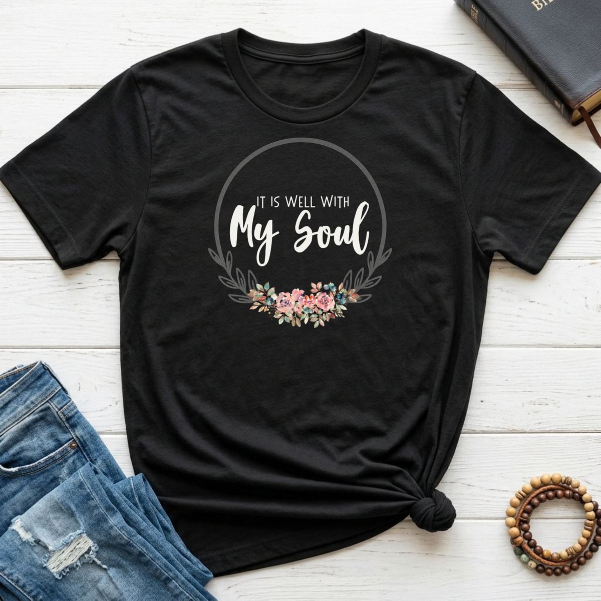 A black short-sleeve graphic T-shirt laid flat on a white wood background, styled with blue jeans, a book, and a beaded bracelet. The shirt has a circular floral print with the text 'It Is Well With My Soul.