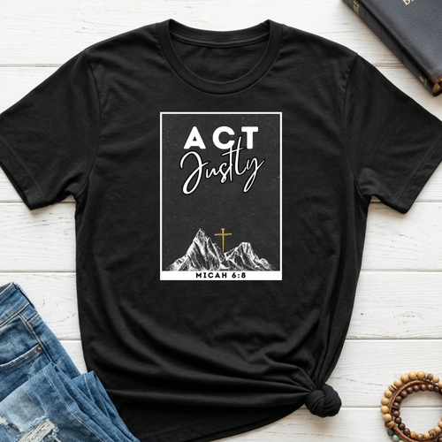 A black short-sleeve graphic T-shirt laid flat on a white wood surface, styled with blue jeans, a book, and a beaded bracelet. The shirt has a white rectangular chest print reading “ACT Justly” with mountain artwork and “MICAH 6:8” below, plus a small gold cross detail.