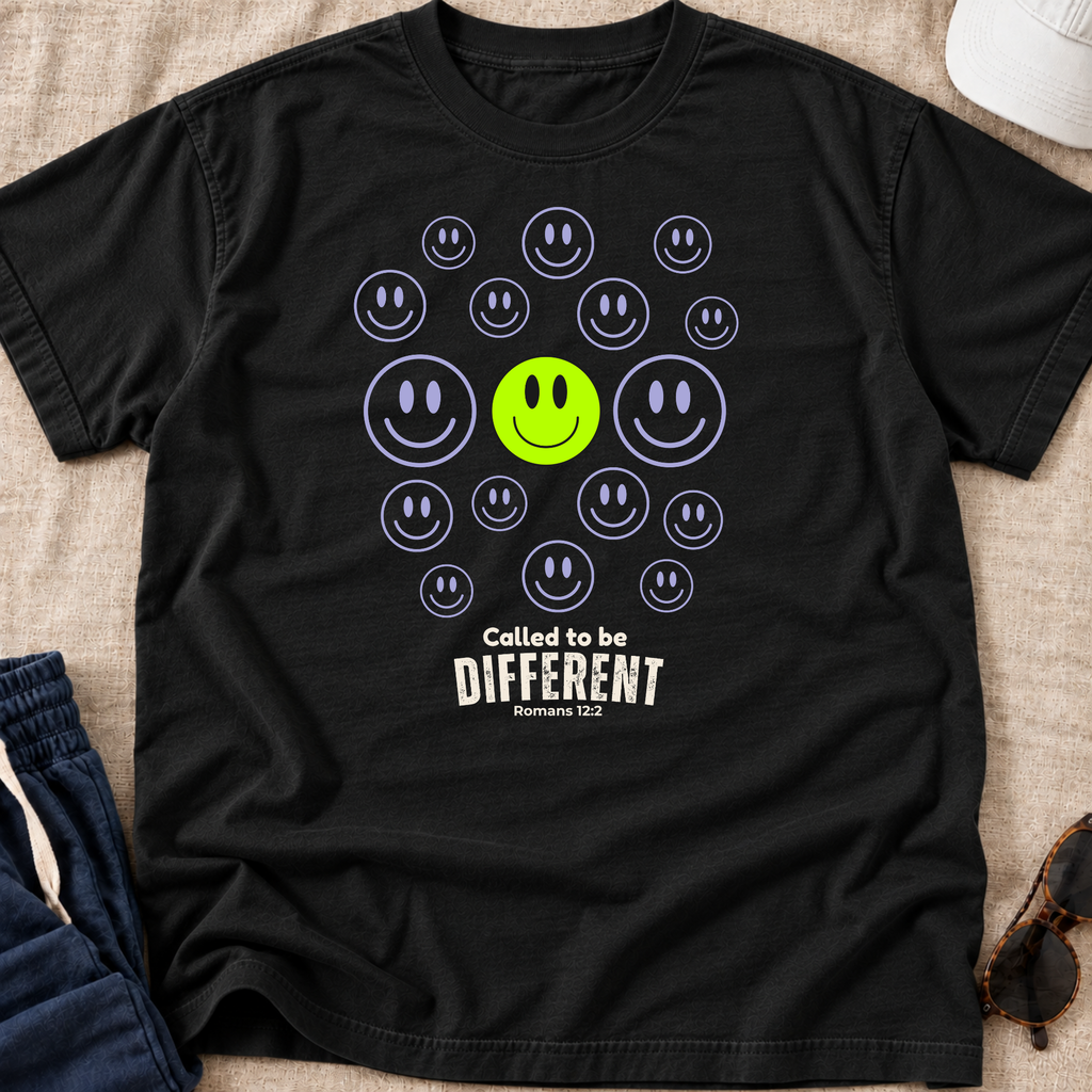 A black short-sleeve graphic T-shirt laid flat on a beige fabric surface, featuring a central smiley-face design with a bright green smiley in the middle surrounded by smaller outlined smileys, and the text 'Called to be DIFFERENT' with 'Numbers 142' below. Partial jeans, a white cap, and sunglasses are visible around the shirt as styling props.