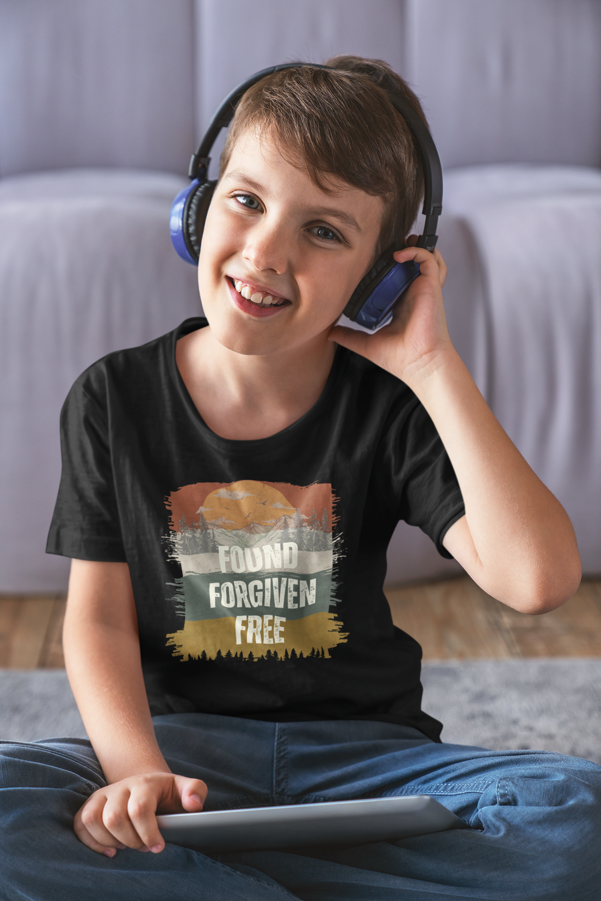 A smiling boy wearing blue over-ear headphones and a black youth Christian graphic T-shirt sits on a rug in a living room setting. The shirt features a distressed sunset graphic with the words 'FOUND FORGIVEN FREE'.