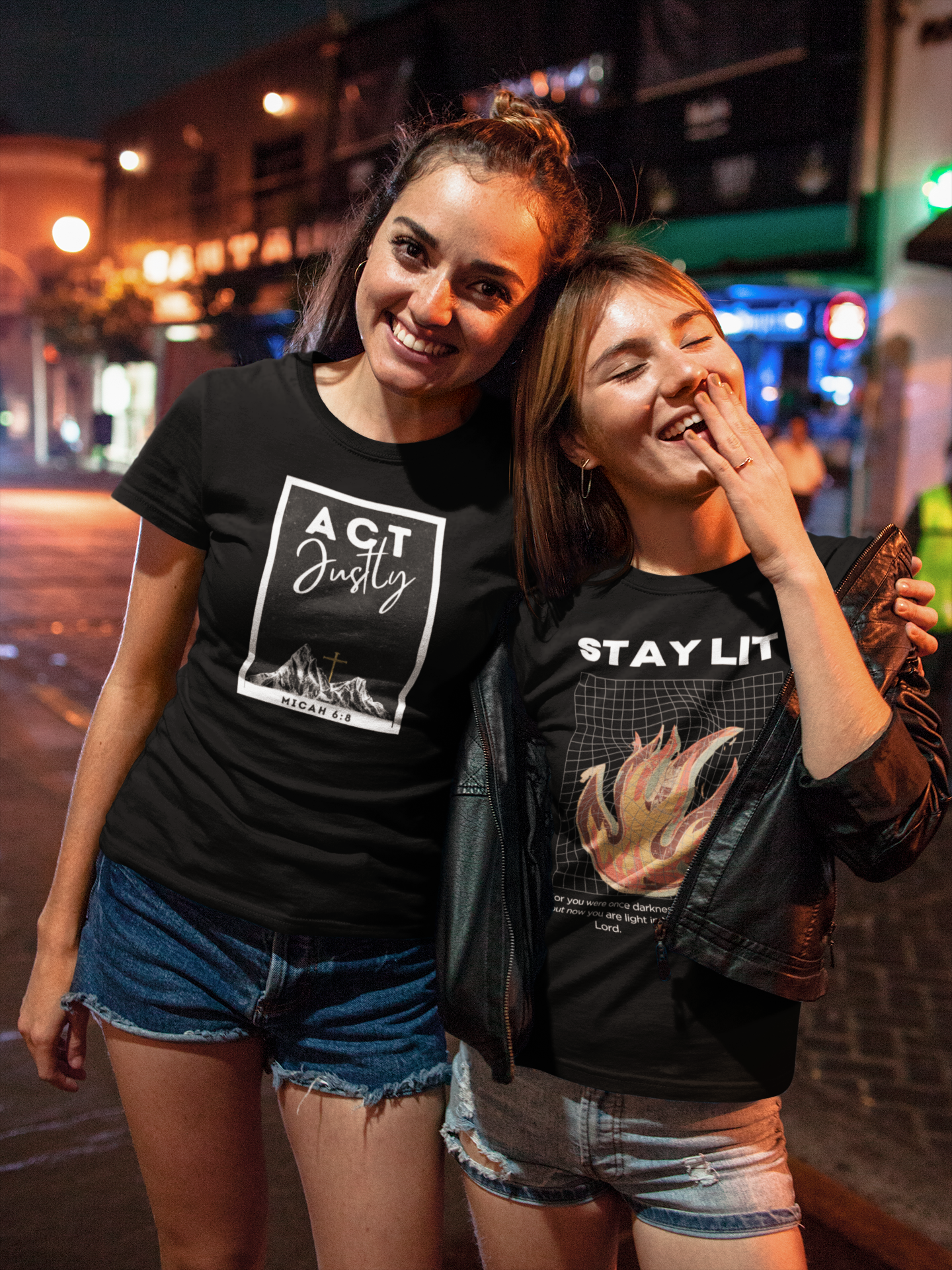 Two women wearing black Christian t-shirts  that say 'act justly' on one, and ' Stay Lit' on the other, with visible text and graphics on a street at night.