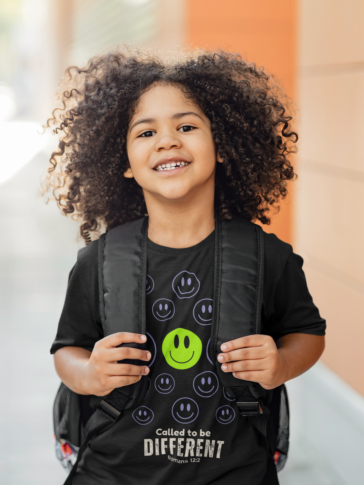 A smiling young girl with curly hair wearing a black graphic t-shirt and a black backpack. The t-shirt features a pattern of smiley faces, with one green smiley face in the center and the text 'Called to be DIFFERENT' printed at the bottom.