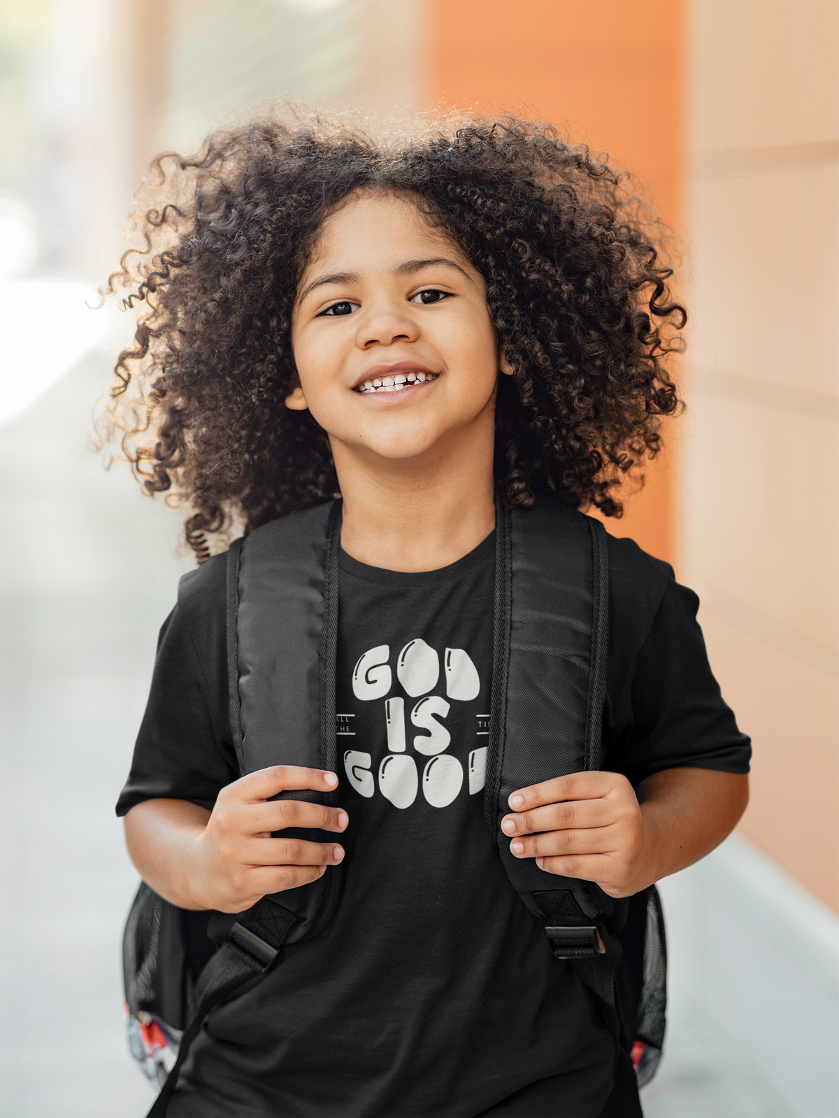 A smiling young kid with voluminous curly hair wears a black Christian graphic t-shirt with 'GOD IS GOOD' printed in white, carrying a black backpack. She is photographed outdoors against a softly blurred background of warm orange and beige tones.