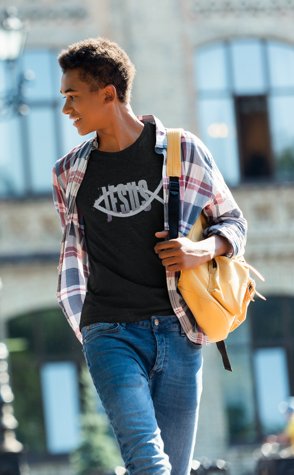 A young man walking outdoors wearing a black Christian graphic t-shirt by SeedSowerGraphics with a white fish symbol and the word 'JESUS', layered under an open red and blue plaid flannel shirt, paired with blue jeans and carrying a yellow backpack.