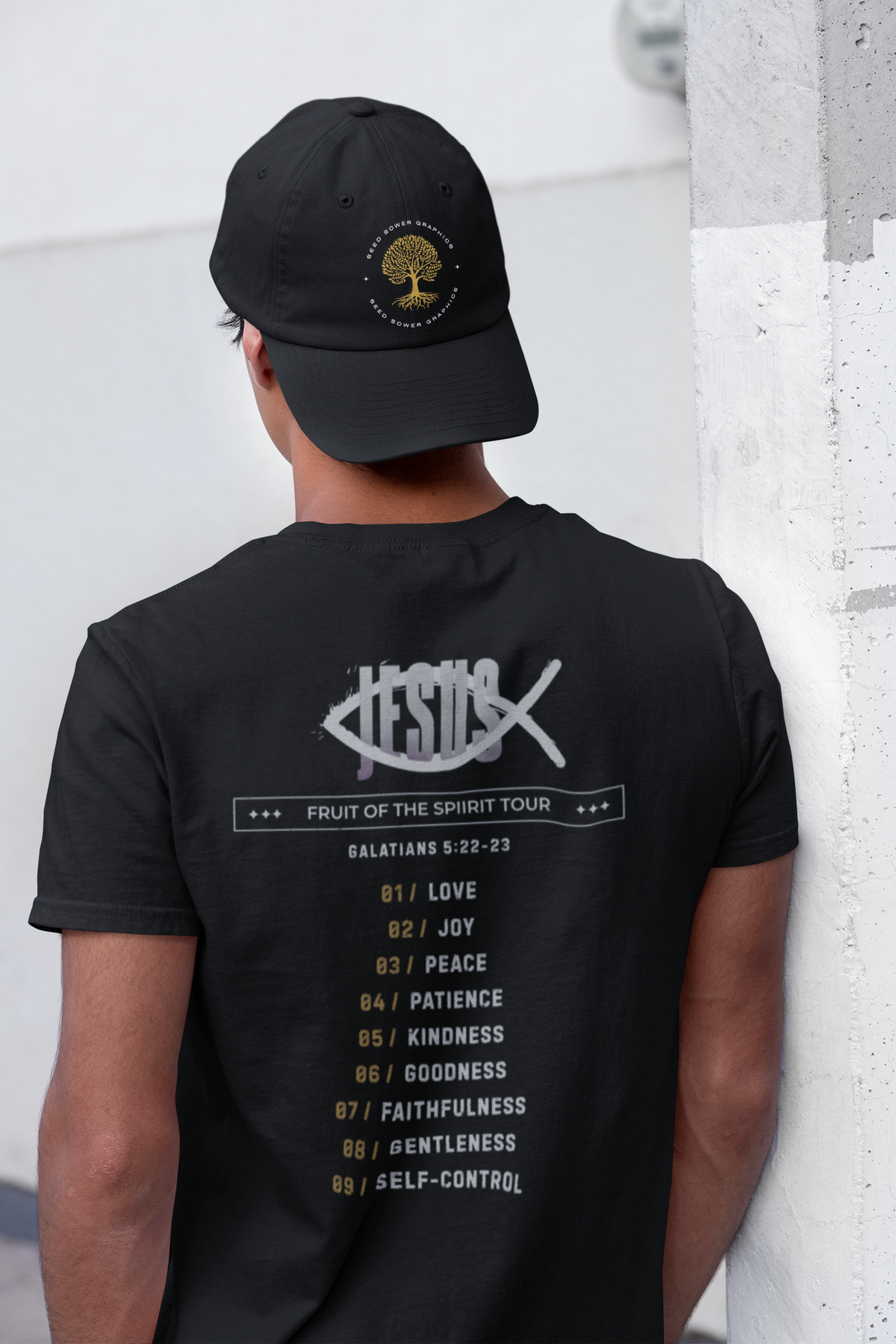 A man wearing a black Chrsitian graphic t-shirt by SeedSowerGraphis and black baseball cap, viewed from behind. The back of the t-shirt features a white graphic of the word 'JESUS' inside an ichthys with the text 'FRUIT OF THE SPIRIT TOUR' and a list of biblical virtues from Galatians 5:22-23.