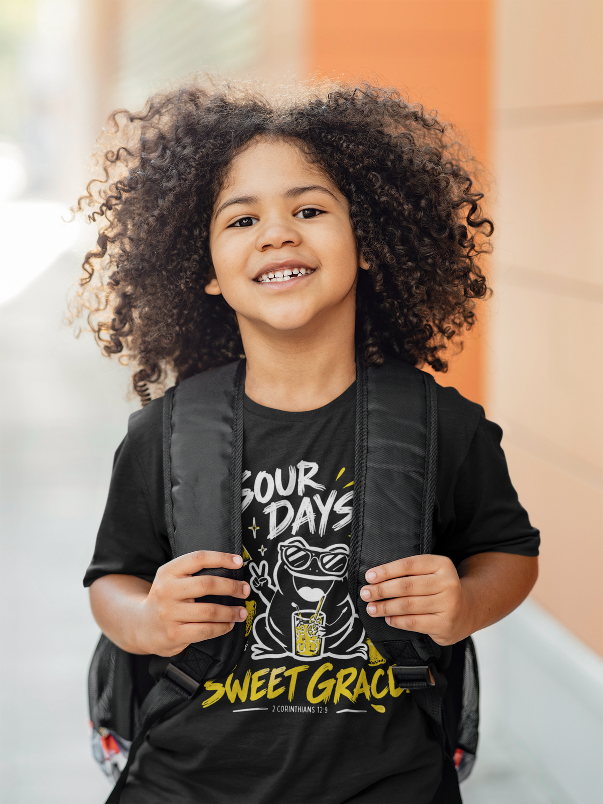 A smiling young kid with voluminous curly hair is shown from the chest up, wearing a black Christian graphic T-shirt by SeedSowerGraphics and a black backpack. The shirt features a cartoon frog wearing sunglasses and the text 'SOUR DAYS SWEET GRACE'.