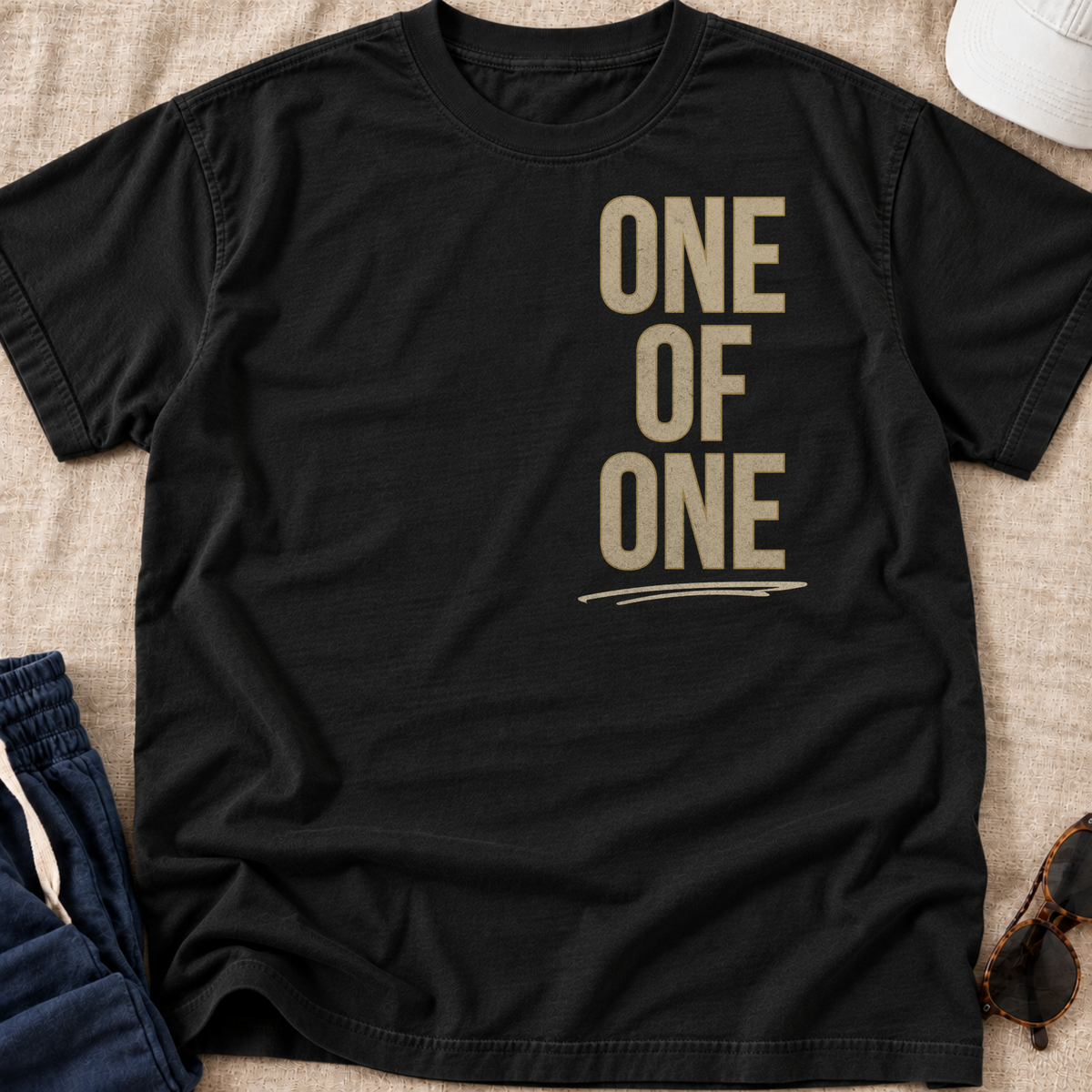 A black short-sleeve t-shirt with the words 'ONE OF ONE' printed in a bold tan serif font on the front, laid flat on a beige textured fabric background with partial styling props visible at the edges.