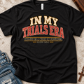 Black christian t-shirt by SeedSowerGraphics  laid out amongst props with typography that says "in my trials era' the joy is a work in progress. James 1:2-4 in cream, red, and black lettering