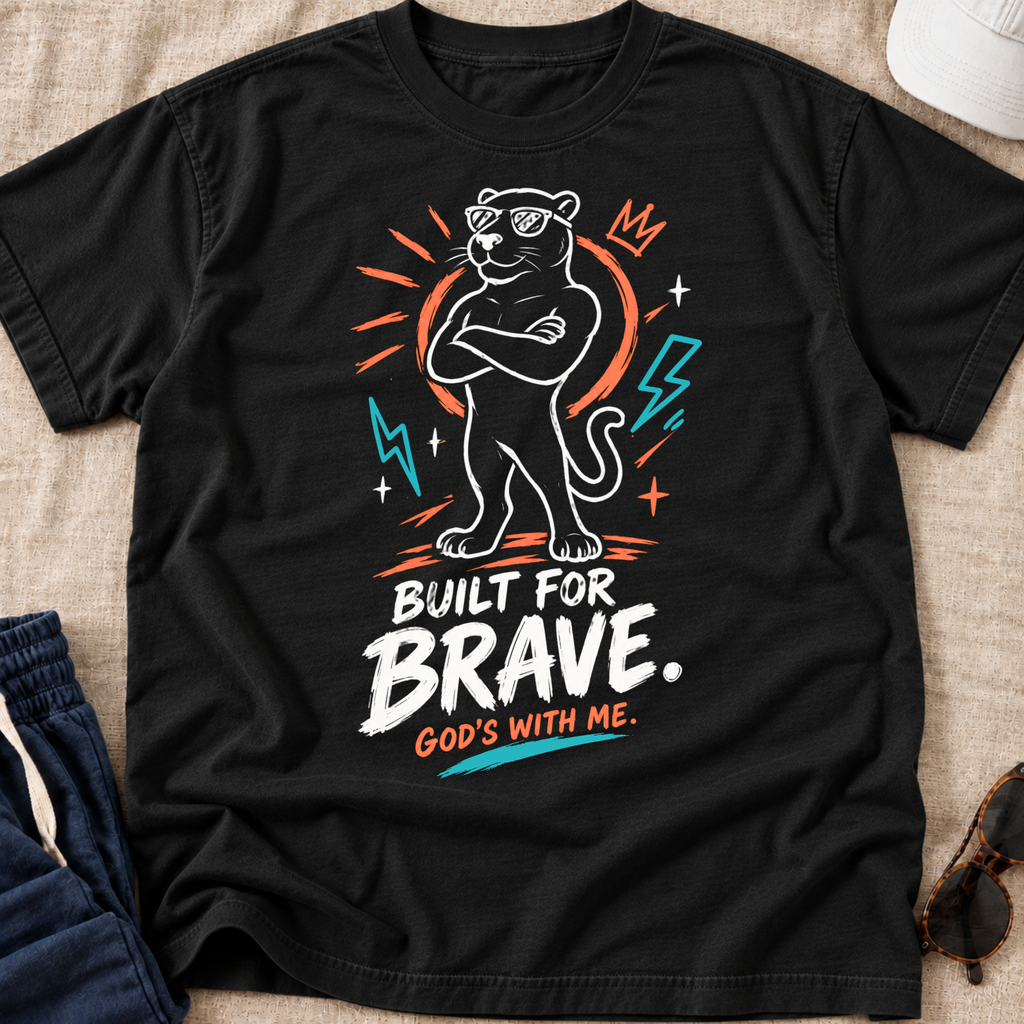 A black graphic t-shirt laid flat on a beige fabric background, featuring a stylized panther illustration with sunglasses, lightning bolts, and the text 'BUILT FOR BRAVE. GOD'S WITH ME.