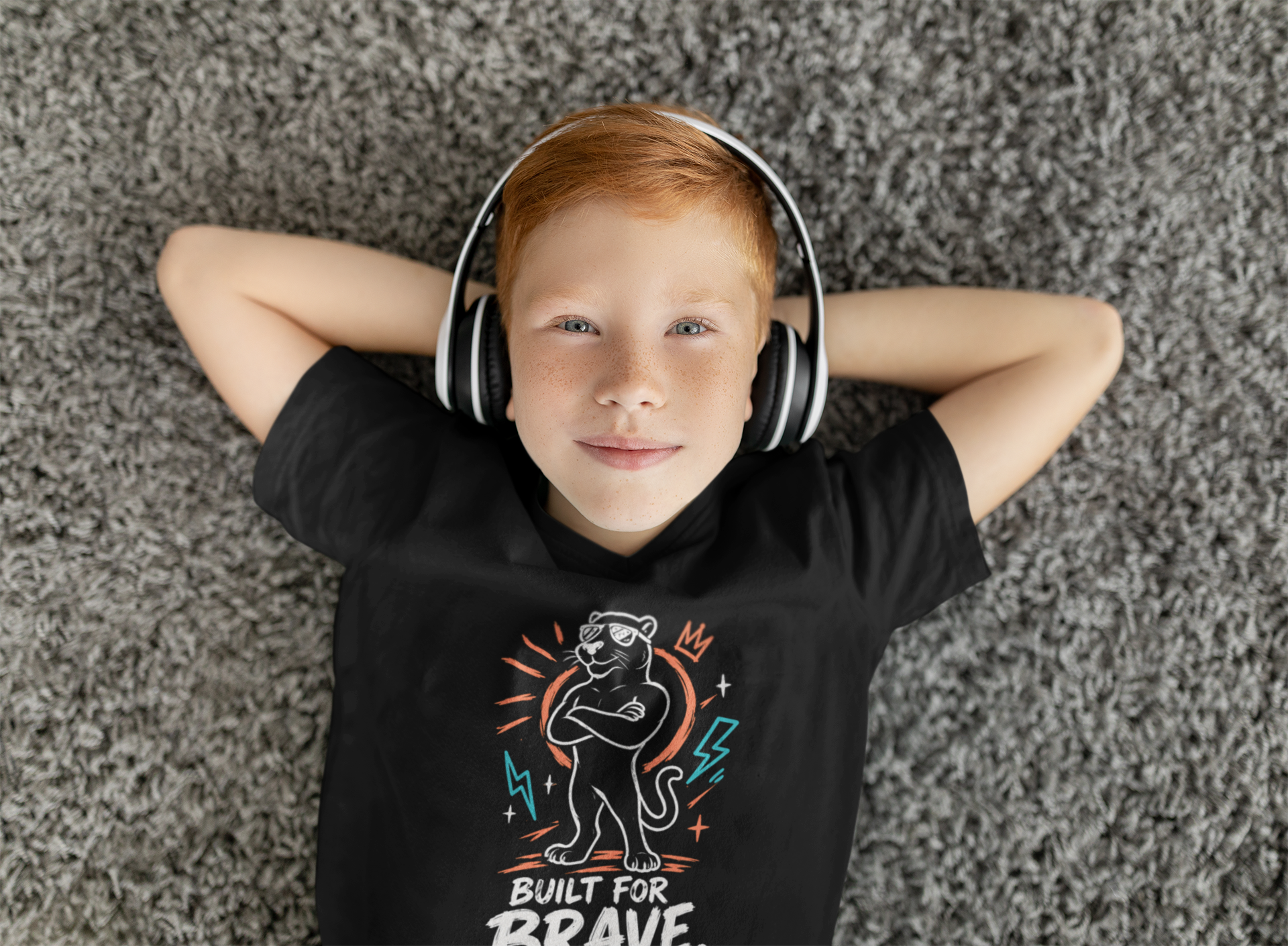 A young boy with red hair lies on a gray textured rug, wearing white over-ear headphones and a black Christian graphic t-shirt by SeedSowerGraphics with a cartoon tiger and the text 'BUILT FOR BRAVE'.