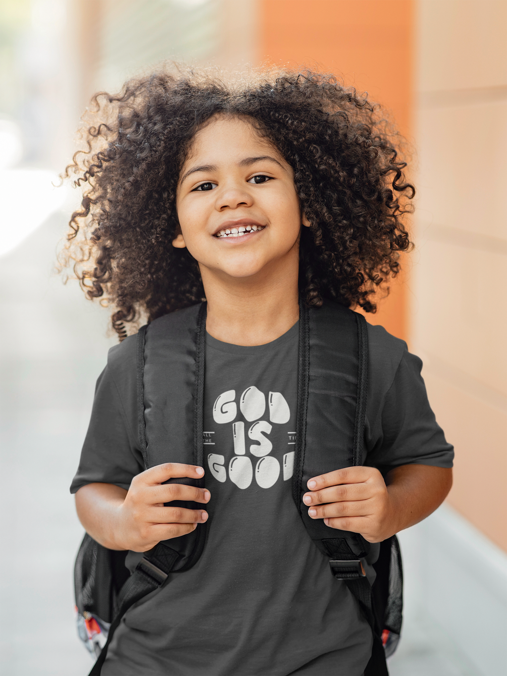 A smiling young girl with voluminous curly hair stands outdoors wearing a dark gray graphic t-shirt that reads 'GOD IS GOOD' in white bubble letters, a black backpack, and looking directly at the camera.