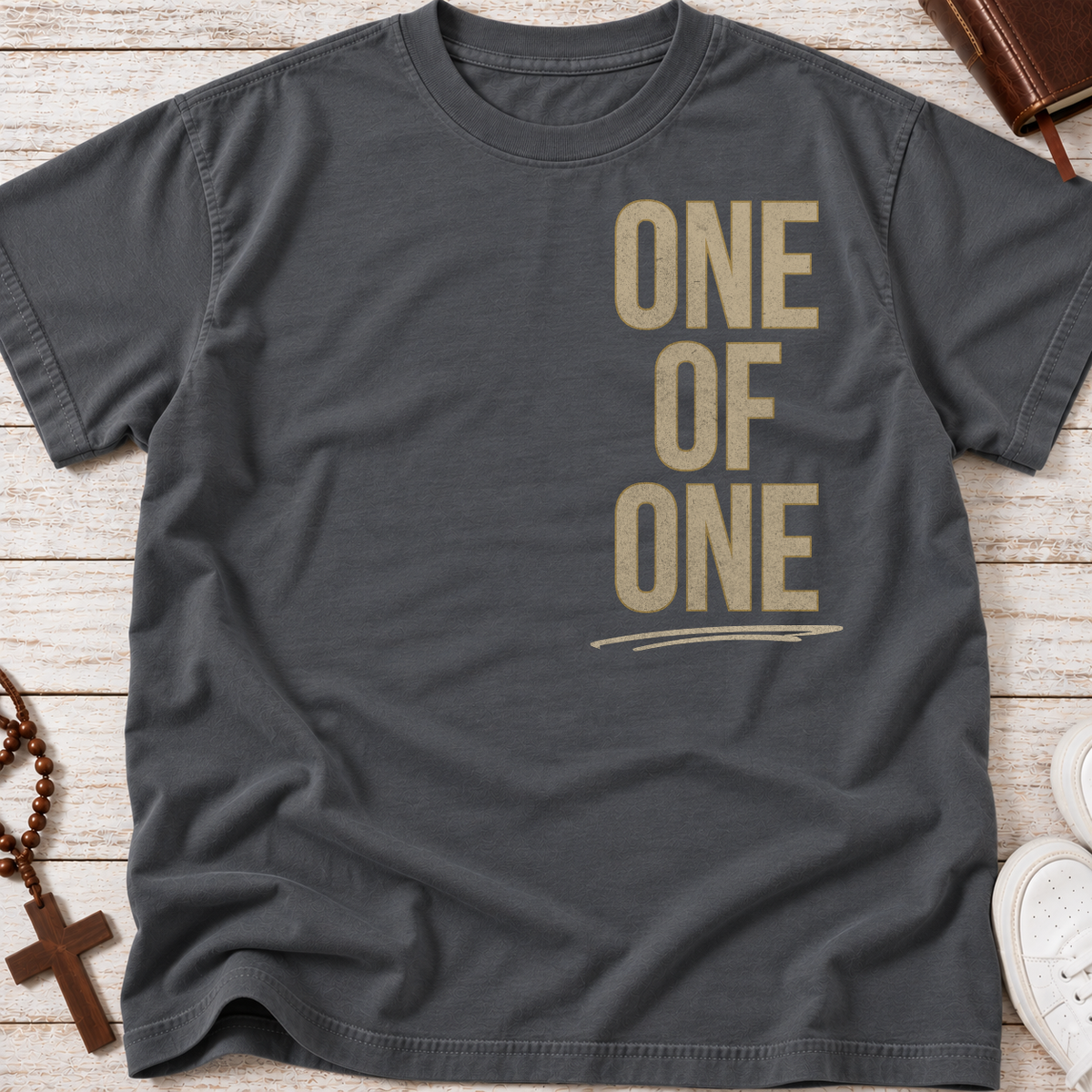 A dark charcoal gray t-shirt with the words 'ONE OF ONE' printed vertically in a distressed tan font, laid flat on a light wood background with lifestyle props like a rosary, cross, and sneakers.
