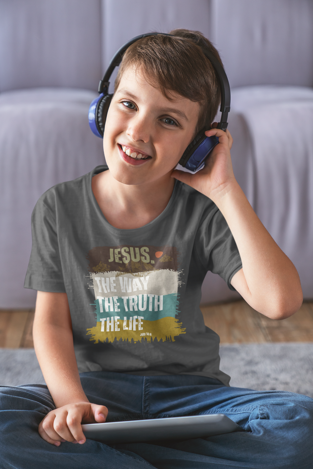A young boy wearing blue over-ear headphones and a gray graphic Christianity t-shirt sits on a rug in front of a bed, holding a tablet. The Christian youth t-shirt features a graphic with the text 'JESUS, THE WAY THE TRUTH THE LIFE' in white, yellow, and brown colors.
