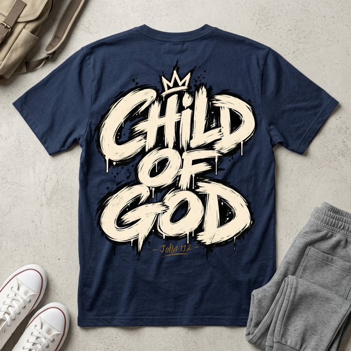 A navy blue t-shirt with a large cream-colored 'CHILD OF GOD' graphic and a gold crown, styled in a flat lay with gray sweatpants, white sneakers, and a beige backpack on a concrete-textured background.