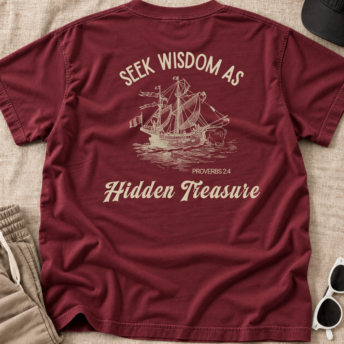 A maroon short-sleeve t-shirt laid flat on a textured beige fabric background, featuring a cream-colored graphic of a sailing ship and the text 'SEEK WISDOM AS Hidden Treasure PROVERBS 2:4'. Partial props including shorts, a hat, and sunglasses are visible around the edges.
