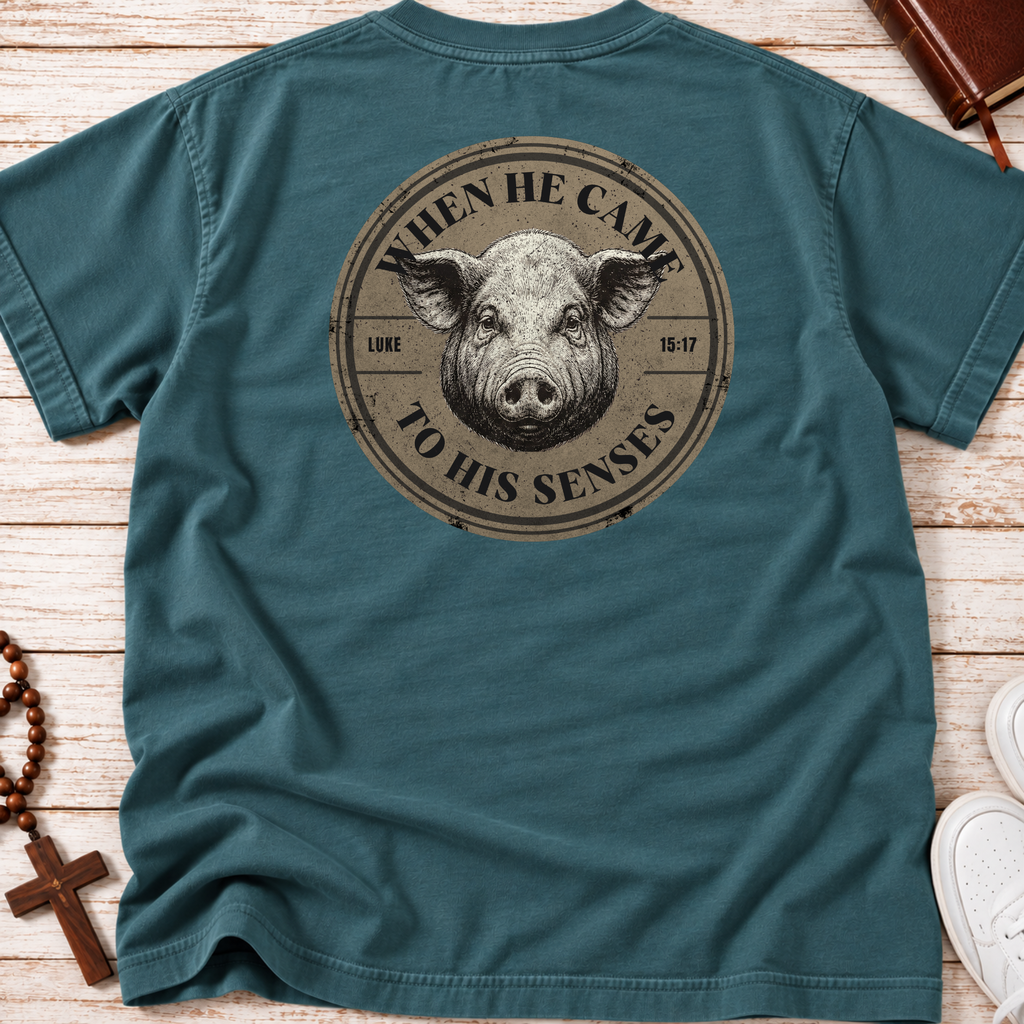 A teal blue short-sleeve t-shirt laid flat on a light wood background, featuring a circular graphic print of a pig's face with the text 'WHEN HE CAME TO HIS SENSES' and 'LUKE 15-17'.