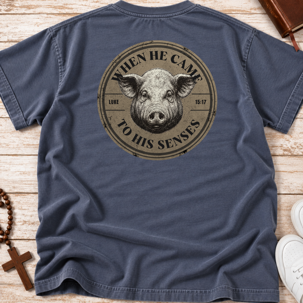A navy blue short-sleeve Christian graphic t-shirt laid flat on a light wood background, featuring a large circular graphic on the back with a pig illustration and the text 'WHEN HE CAME TO HIS SENSES' and 'LUKE 15-17'.