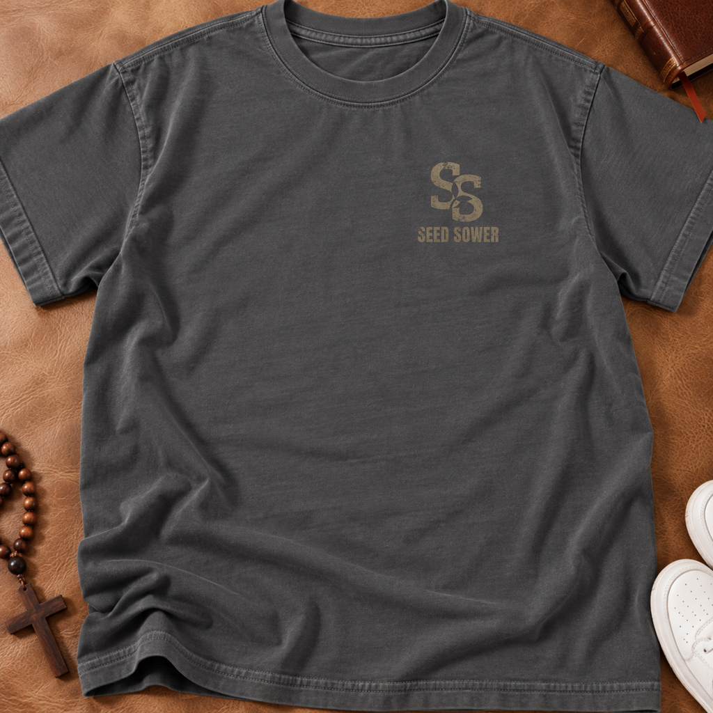 A dark charcoal gray crewneck t-shirt with a distressed tan 'SS SEED SOWER' logo on the left chest, laid flat on a brown leather background with styled props including a wooden cross, beads, and a book.
