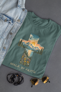 A sage green comfort colors Christian graphic t-shirt featuring a cross-shaped landscape print with a sunset and the text 'WALK BY FAITH' and '2 CORINTHIANS 5:7', styled in a flat lay with a denim jacket, sunglasses, and beaded bracelets on a gray background.