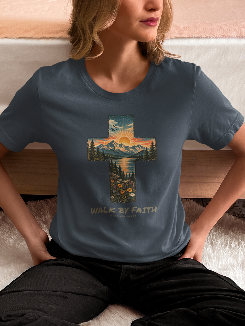 A woman wearing a slate blue Faith cross crewneck t-shirt with a large cross graphic containing a mountain sunset landscape and the text 'WALK BY FAITH' printed below it, sitting on a white rug in front of a bed.