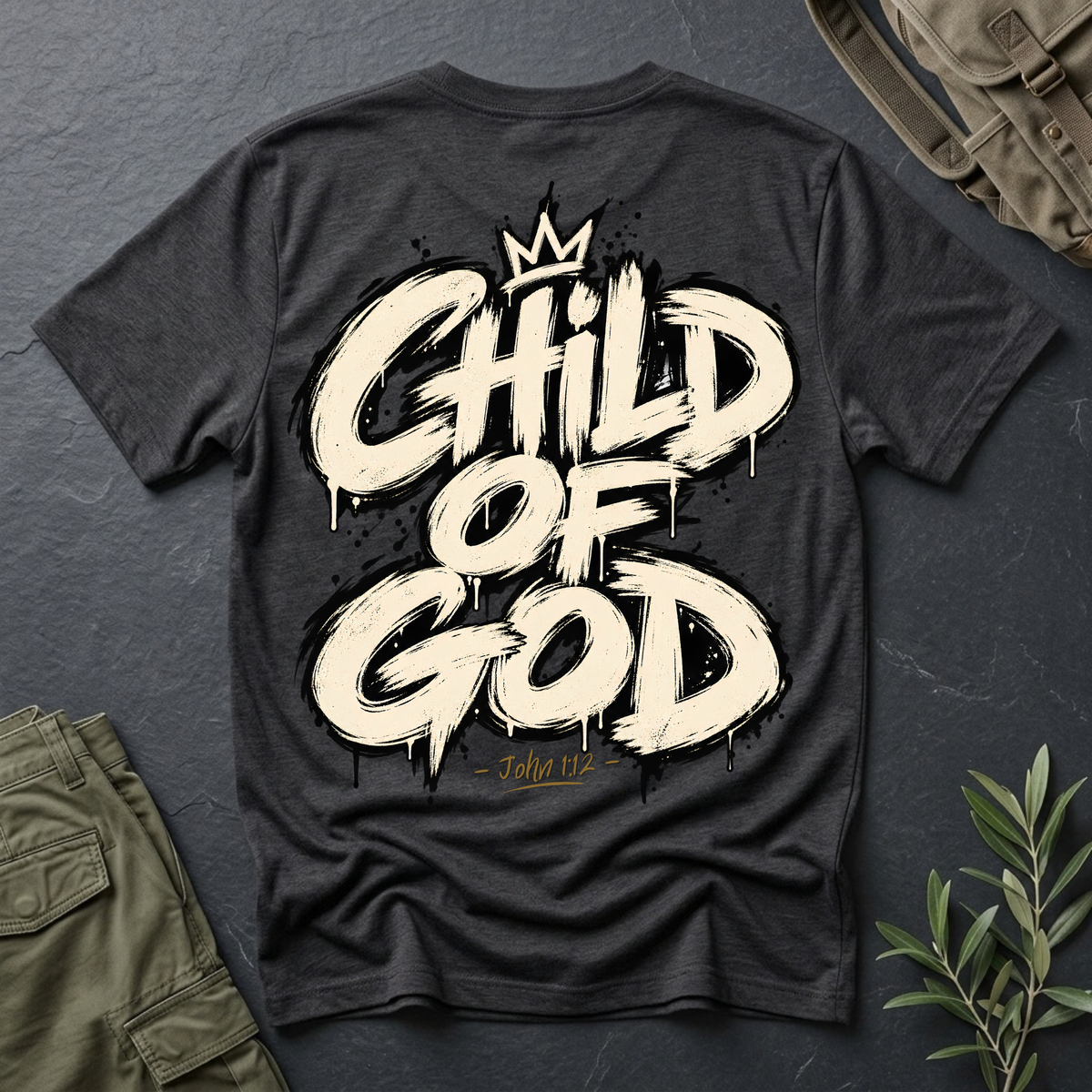 A dark charcoal gray t-shirt shown from the back, featuring a large cream-colored graphic that reads 'CHILD OF GOD' with a small crown above and 'John 1:12' below in gold. The shirt is styled in a flat-lay arrangement on a dark slate background with an olive green backpack, cargo pants, and a small green plant visible.