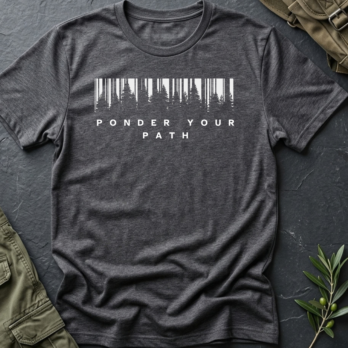 A dark heather short-sleeve t-shirt laid flat on a textured beige surface, featuring a white graphic of a forest silhouette inside a barcode shape above the words 'PONDER YOUR PATH'. Blue jeans are partially visible at the bottom left, and a wooden cross is at the bottom right.