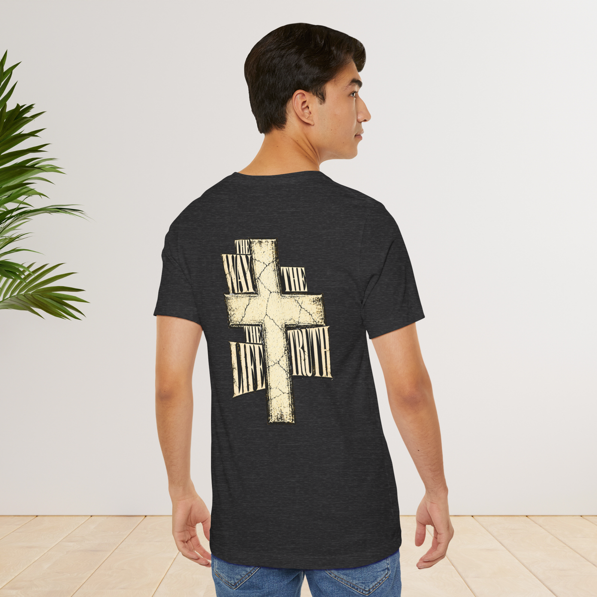 A man wearing a dark heather gray Christian graphic t-shirt by SeedSowerGraphics with a large cream-colored cross graphic on the back, standing in a room with a light wall and wood floor.