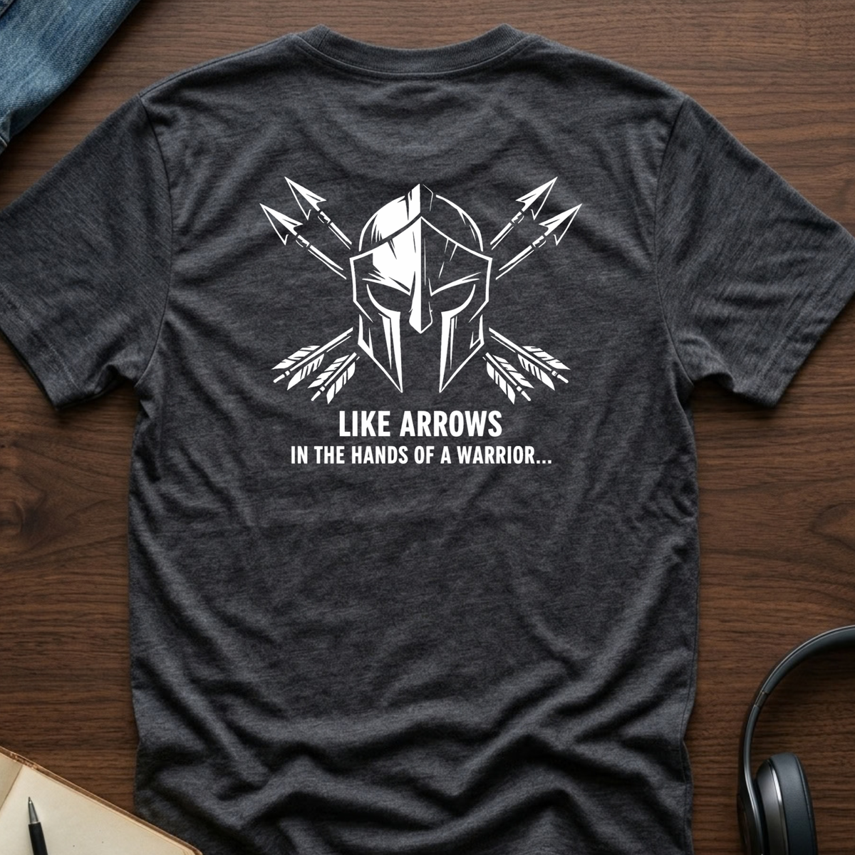 A dark heather gray t-shirt laid flat on a wooden surface, featuring a white graphic of a Spartan helmet with crossed arrows and the text 'LIKE ARROWS IN THE HANDS OF A WARRIOR.