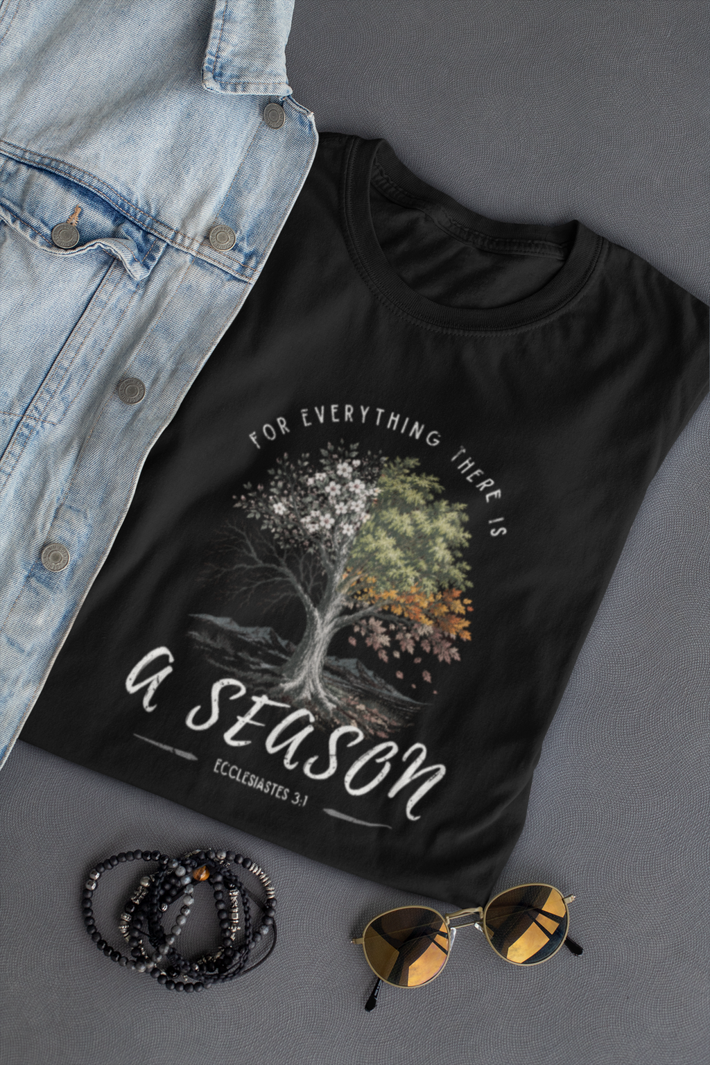 A black Christian graphic T-shirt by SeedSowerGraphics laid flat on a gray surface, styled with a light blue denim jacket, beaded bracelets, and round sunglasses. The shirt features a tree illustration with white, green, and orange foliage and the text 'FOR EVERYTHING THERE IS A SEASON' and 'EXCELELSIastes 3:1'.
