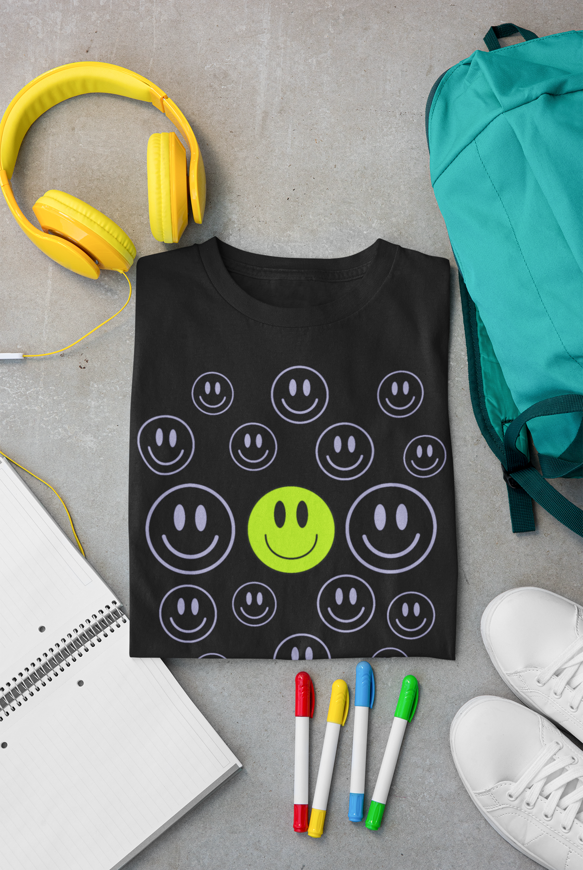A black short-sleeve Christian graphic T-shirt by SeedSowerGraphics laid flat on a gray concrete surface, featuring a pattern of white smiley faces with one central lime green smiley face. The shirt is styled with yellow headphones, a teal backpack, white sneakers, colored markers, and an open notebook around it.