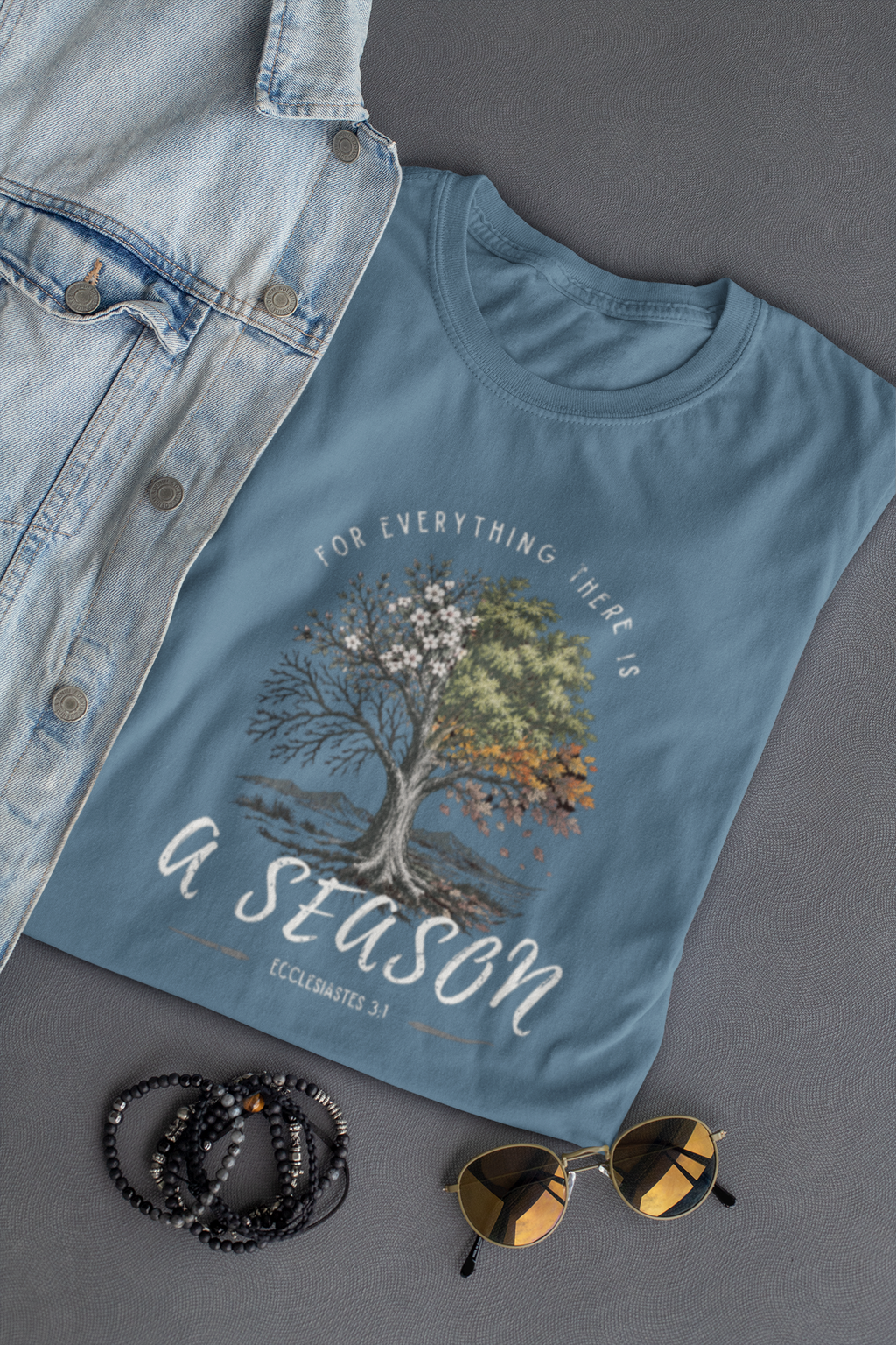 A slate blue Christian graphic t-shirt by SeedSowerGraphics featuring a tree design with white, pink, green, and orange leaves, surrounded by the text 'FOR EVERYTHING THERE IS A SEASON' and 'ECCLESIAstes 3:1'. The shirt is styled in a flat-lay arrangement with a light-wash denim jacket, black beaded bracelets, and gold-framed sunglasses on a dark gray background.