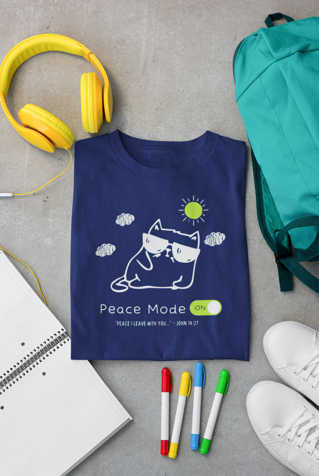A navy blue Christian graphic t-shirt by SeedSowerGraphics featuring a white line art illustration of a cat wearing sunglasses with a sun and clouds, and the text 'Peace Mode ON!'. The shirt is presented in a styled flat-lay arrangement on a gray concrete floor alongside yellow headphones, a teal backpack, a notebook, colored markers, and white sneakers.