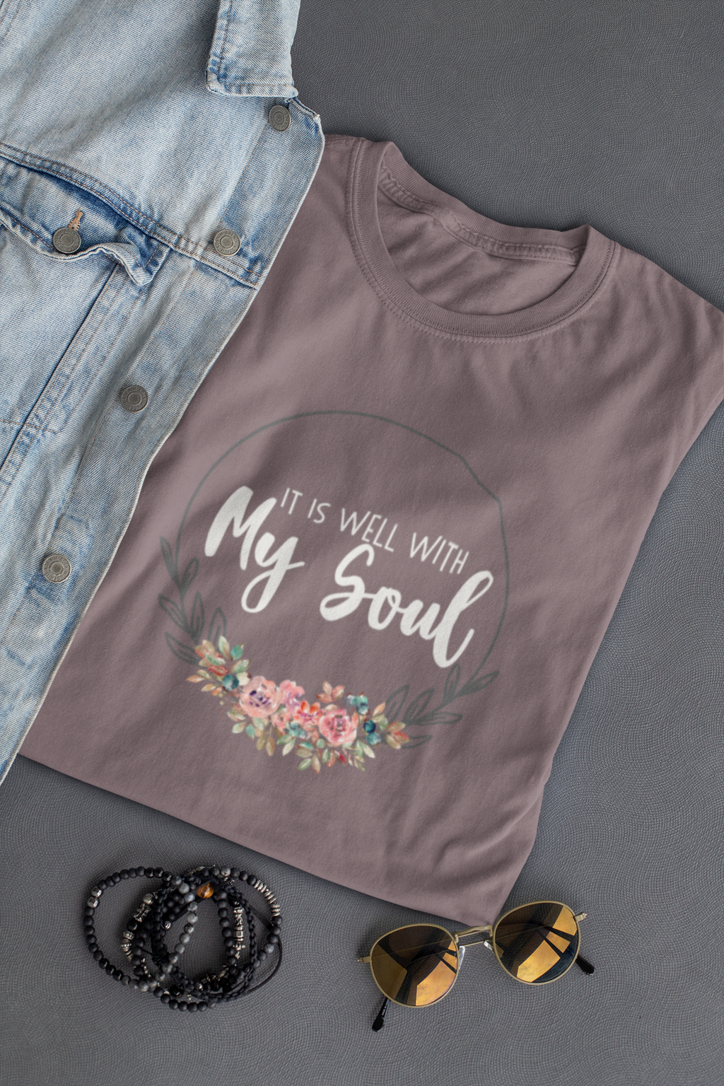 A mauve-gray Christian graphic t-shirt by SeedSowerGraphics with a floral design and the text 'IT IS WELL WITH My Soul' in white, styled in a flat-lay with a denim jacket, beaded bracelets, and sunglasses on a dark gray background.