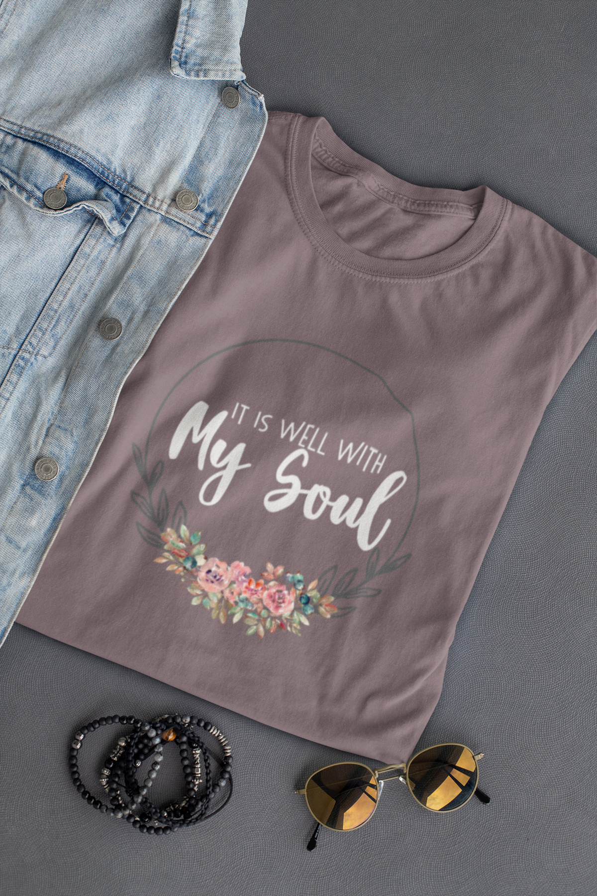 A mauve-gray Christian graphic t-shirt by SeedSowerGraphics with a floral design and the text 'IT IS WELL WITH My Soul' in white, styled in a flat-lay with a denim jacket, beaded bracelets, and sunglasses on a dark gray background.