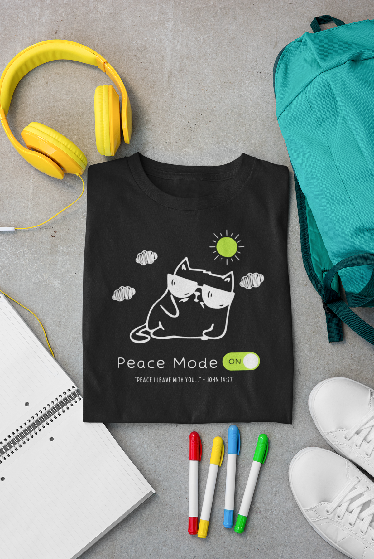 A black Christian graphic t-shirt by SeedSowerGraphics featuring a white line art illustration of a cat wearing sunglasses with a green sun and the text 'Peace Mode ON!', styled in a flat lay on a gray concrete surface with yellow headphones, a teal backpack, a notebook, colored markers, and white sneakers.