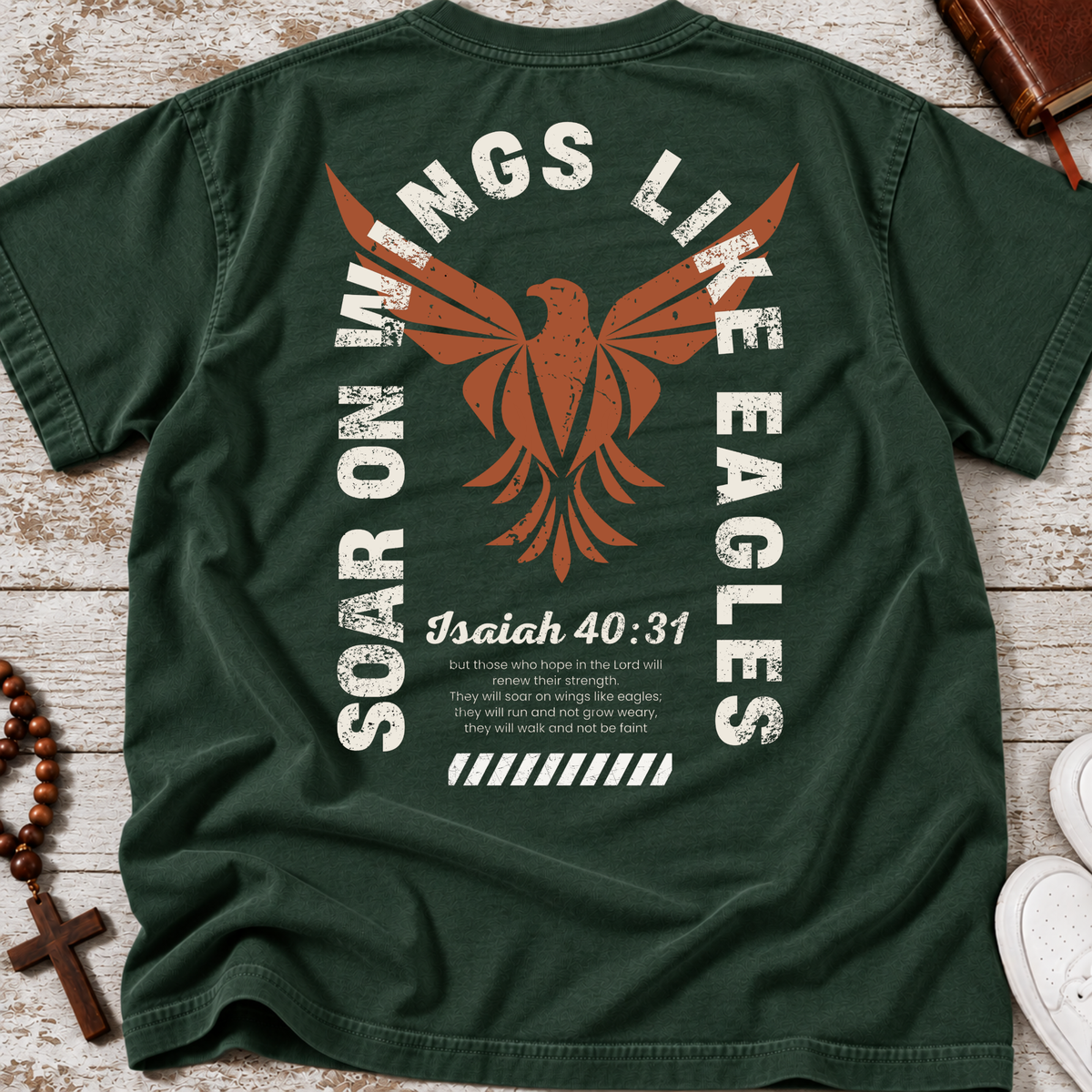 A forest green short-sleeve t-shirt laid flat on a light wood surface, showing the back graphic with a large orange eagle, distressed white text reading 'Soar on Wings Like Eagles' and 'Isaiah 40:31', plus smaller scripture text. A cross necklace and Bible and part of white shoes are visible as styling props.
