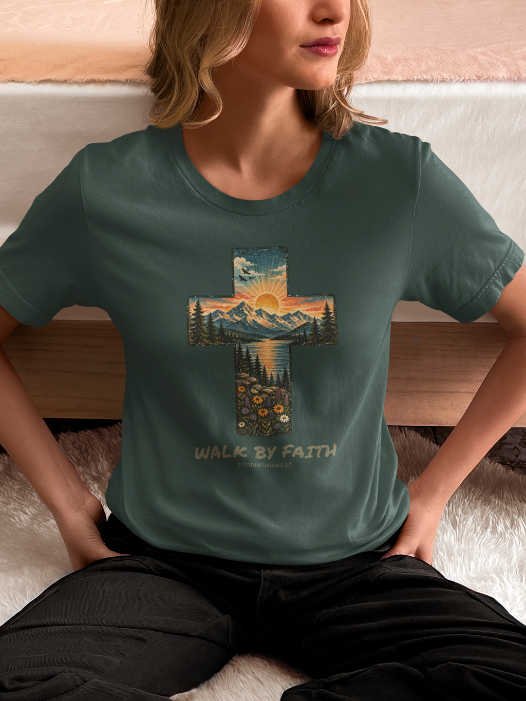 A woman wearing a sage green Bible verse t-shirt with a large cross graphic containing a mountain sunset landscape and the text 'WALK BY FAITH' printed below it, sitting on a white rug in front of a bed.