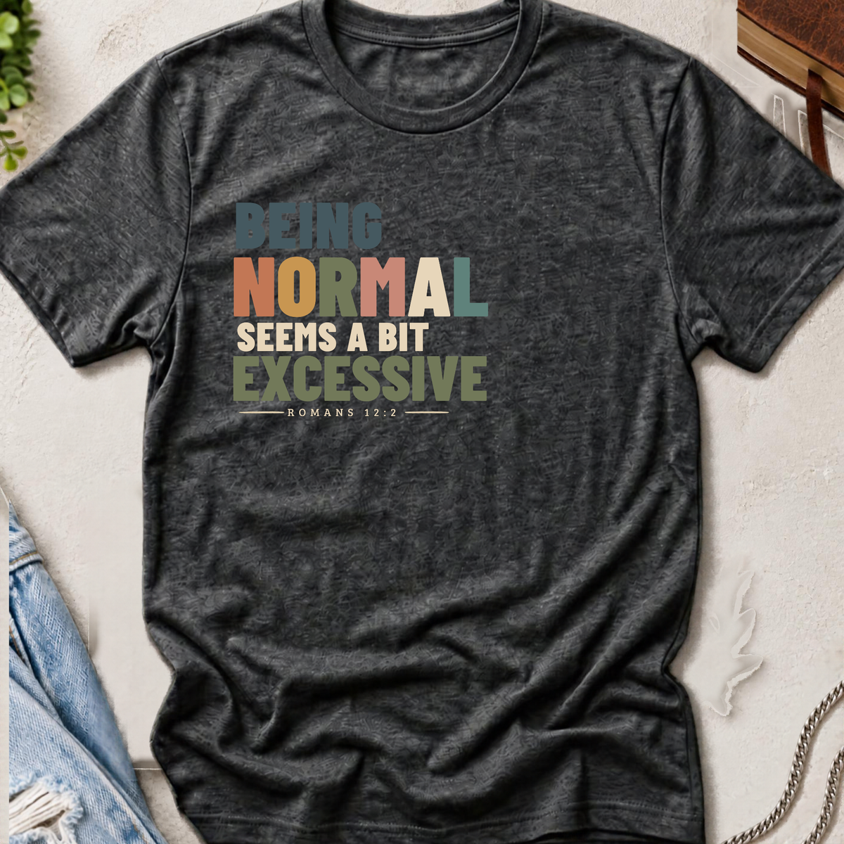 A dark heather gray short-sleeve graphic T-shirt laid flat on a textured light surface with jeans and a wooden cross as styling props. The shirt has a multicolor typographic slogan reading “BEING NORMAL SEEMS A BIT EXCESSIVE” with “ROMANS 13:2” underneath.