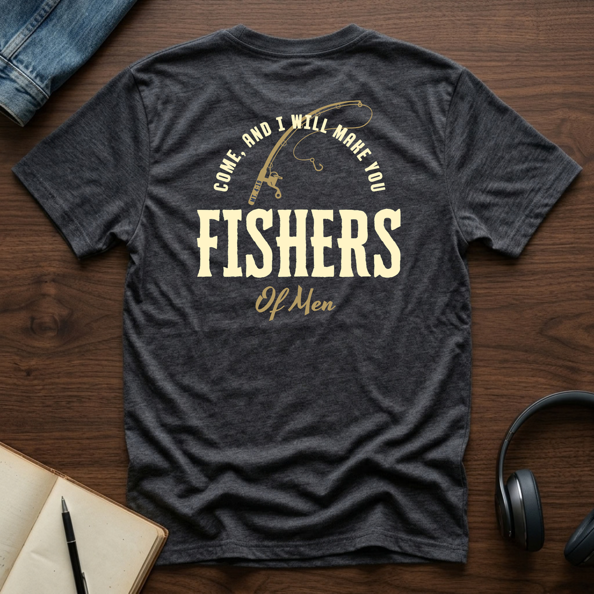 A dark heather gray Christian graphic t-shirt laid flat on a wooden surface, featuring a vintage-style cream graphic that reads 'COME, AND I WILL MAKE YOU FISHERS OF MEN' with a fishing rod illustration. The shirt is styled with a denim jacket, headphones, an open notebook, and a pen.