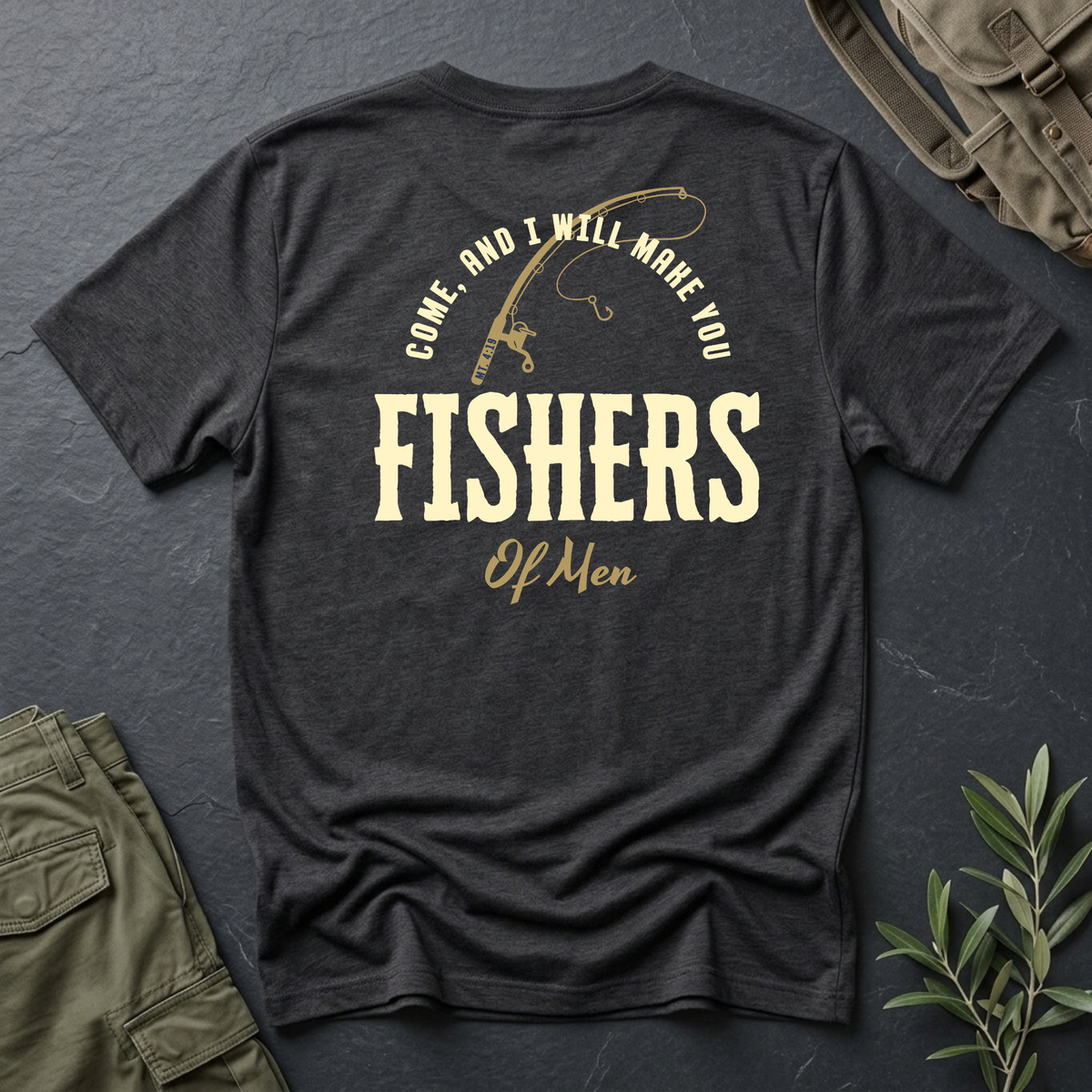 A dark charcoal heather Christian t-shirt with a vintage-style cream graphic on the back reading 'COME AND I WILL MAKE YOU FISHERS OF MEN' with a fishing rod illustration, styled in a flat-lay with olive cargo pants, a backpack, and a small green plant on a dark slate background.
