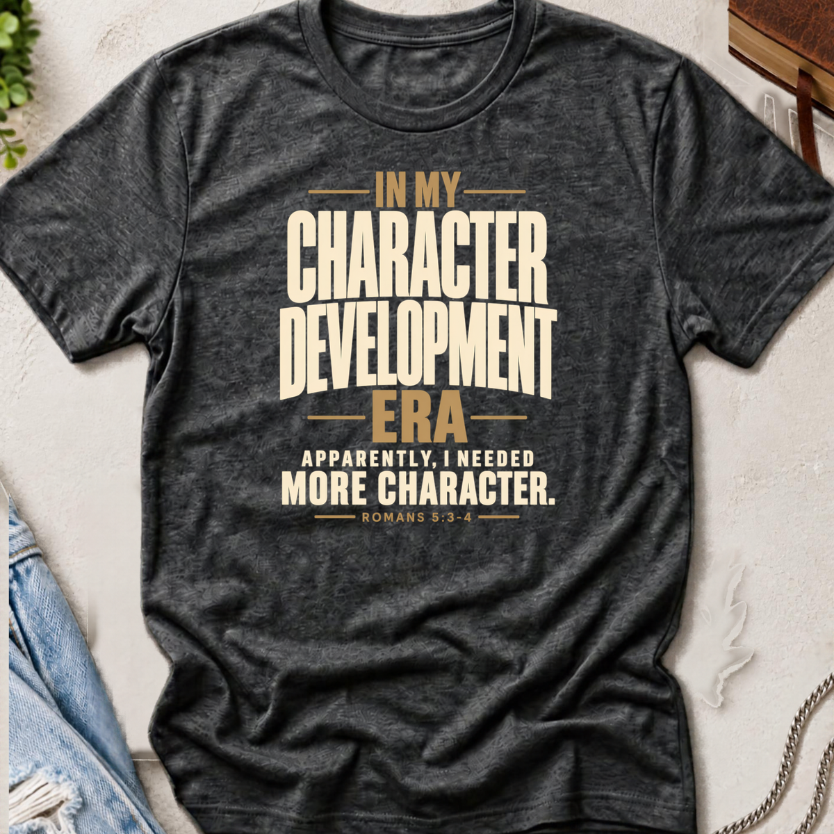 A dark heather gray Christian graphic t-shirt by SeedSowerGraphics laid flat on a textured light background, styled with a denim jacket corner, a plant leaf, and a chain. The t-shirt features a beige and gold typographic design that reads: 'IN MY CHARACTER DEVELOPMENT ERA APPARENTLY, I NEEDED MORE CHARACTER.