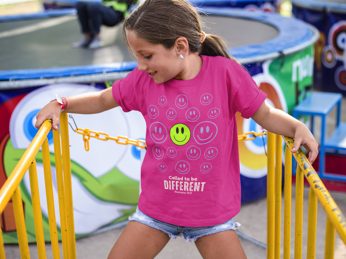 A young girl smiling while wearing a bright pink graphic t-shirt with smiley faces and the text 'Called to be DIFFERENT' on a playground with yellow railings.