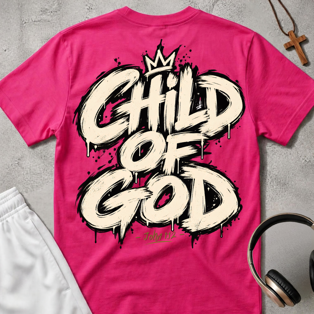 A bright pink t-shirt laid flat on a gray concrete-textured background, featuring a large white and black spray-paint style graphic that reads 'CHILD OF GOD' with a small crown above and 'John 1:12' below. White shorts are partially visible on the left, and a wooden cross necklace and over-ear headphones are placed on the right.