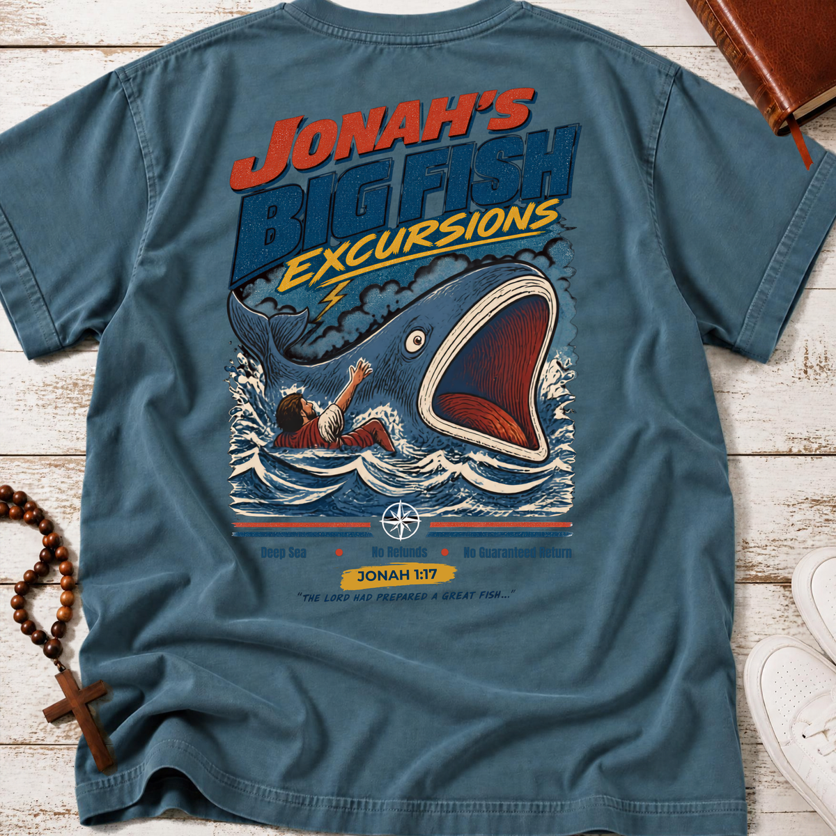 An ice blue Christian graphic t-shirt by SeedSowerGraphics laid flat on a white wood plank background, featuring a large vintage-style illustration of a giant whale and a man on the back. The shirt is styled with a wooden rosary, a brown leather book, and white sneakers.